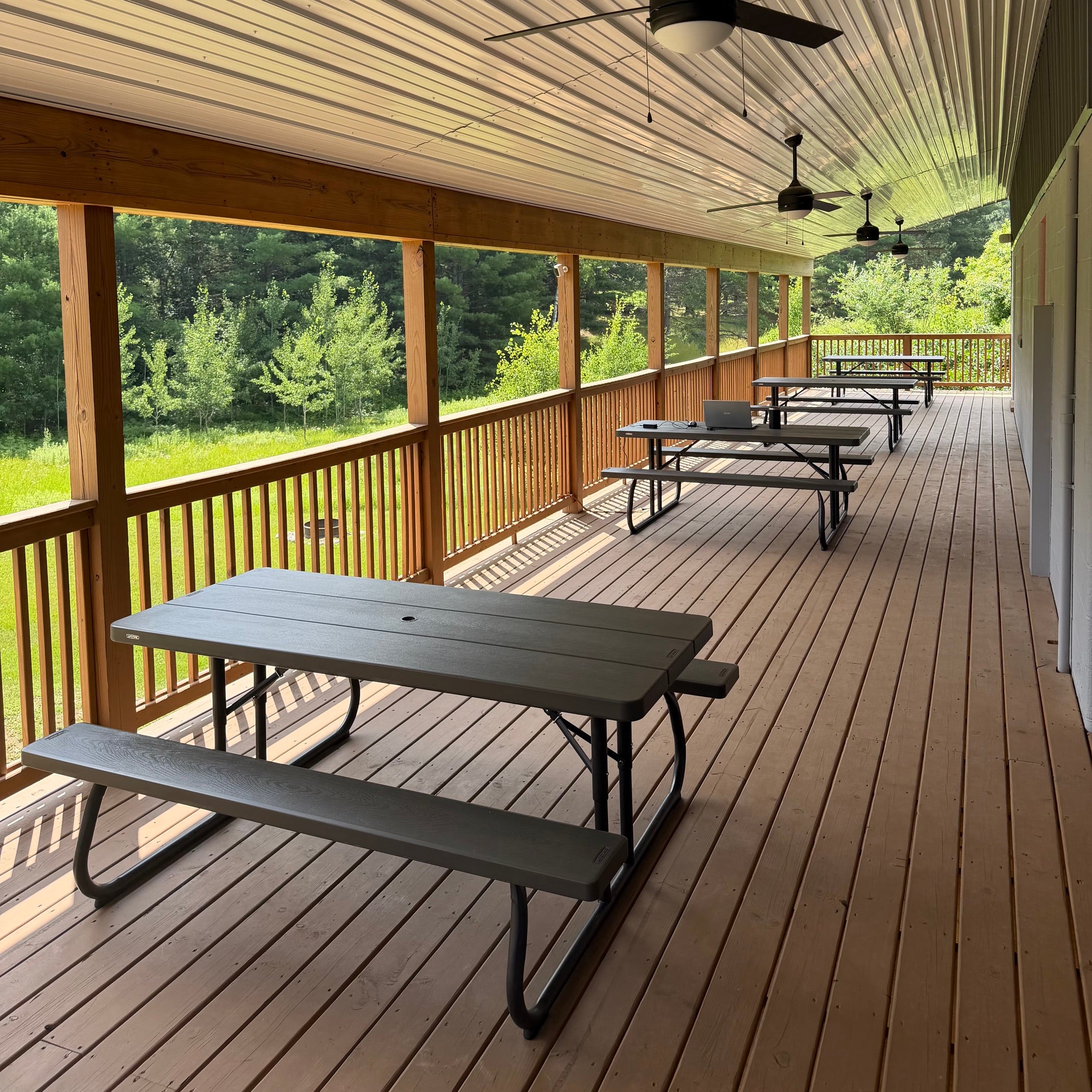 A covered wooden deck with several gray picnic tables, railings, ceiling fans, and a view of green trees outside.