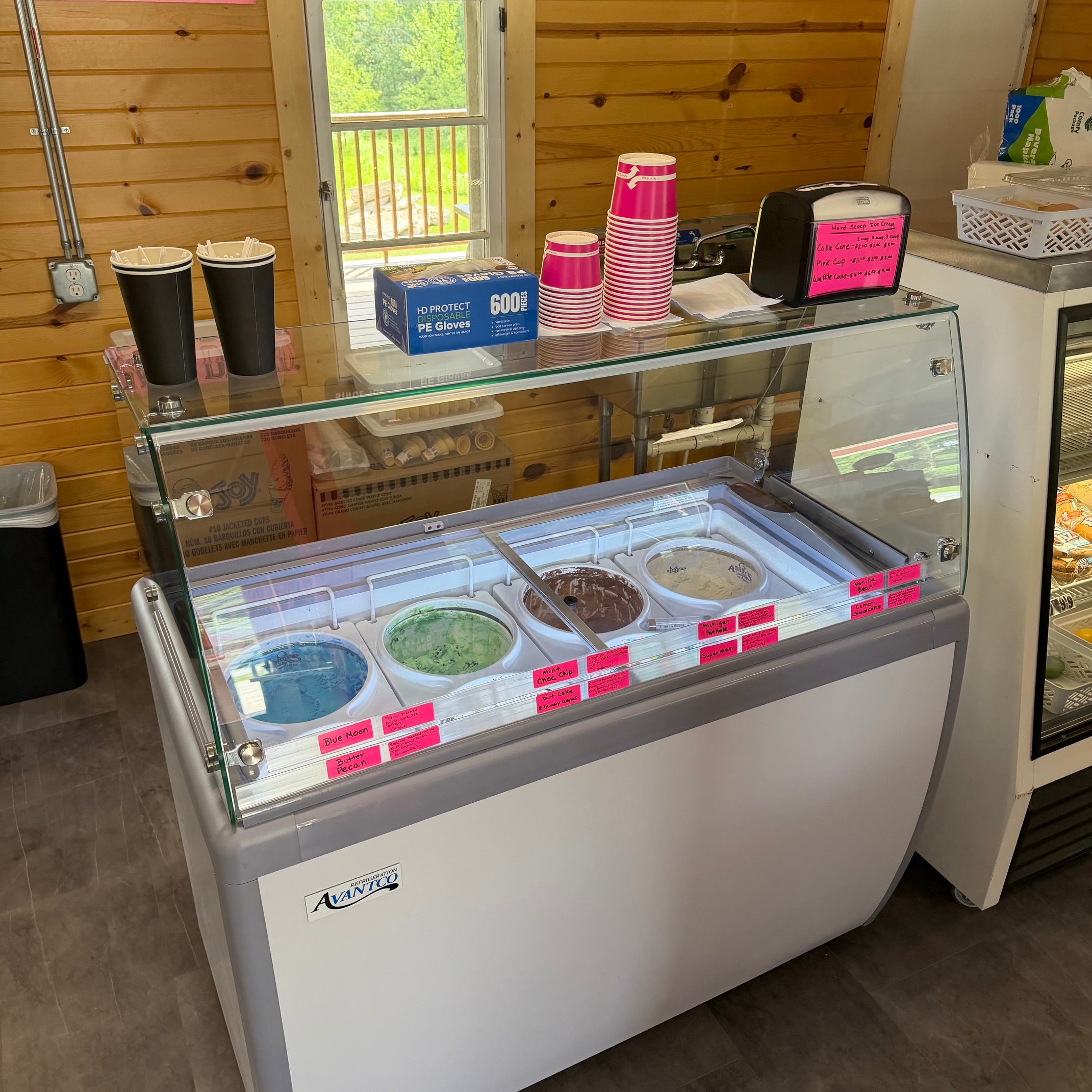 Hard-scoop ice cream freezer with a glass display cover, showing several ice cream flavors. The freezer is set in a wood-paneled shop interior with cups, napkins, and other supplies on top.